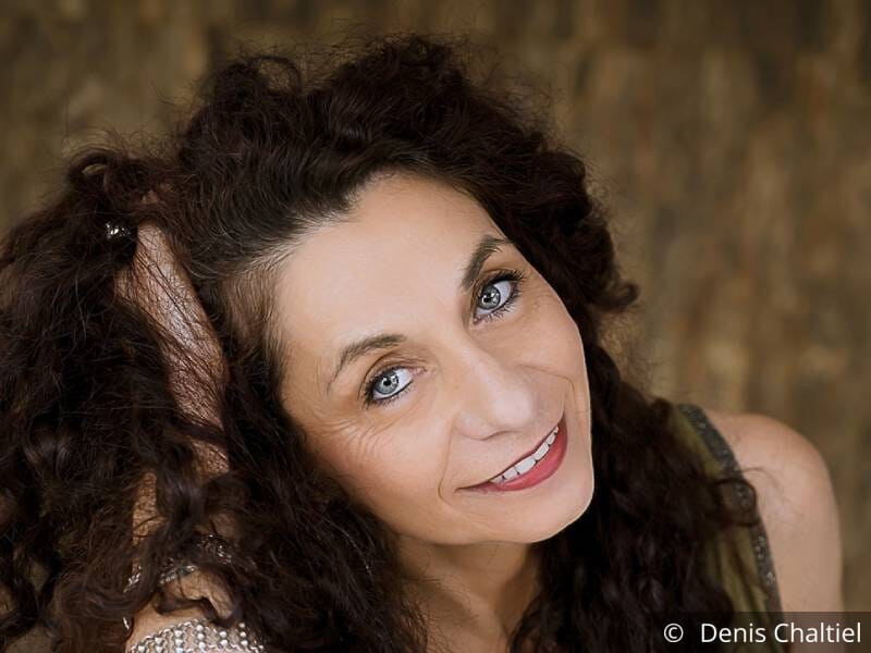 Portrait d'une femme brune aux cheveux ondulés, yeux bleus et qui sourit.